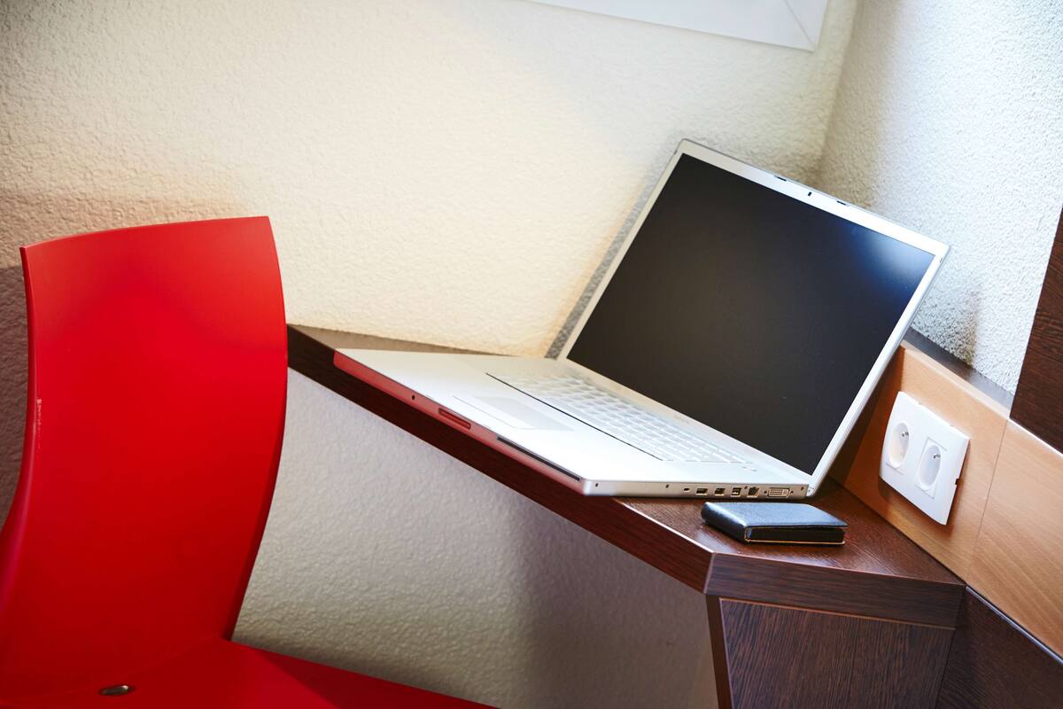 Una computadora portátil sobre un escritorio con una silla roja, un teléfono y un enchufe.