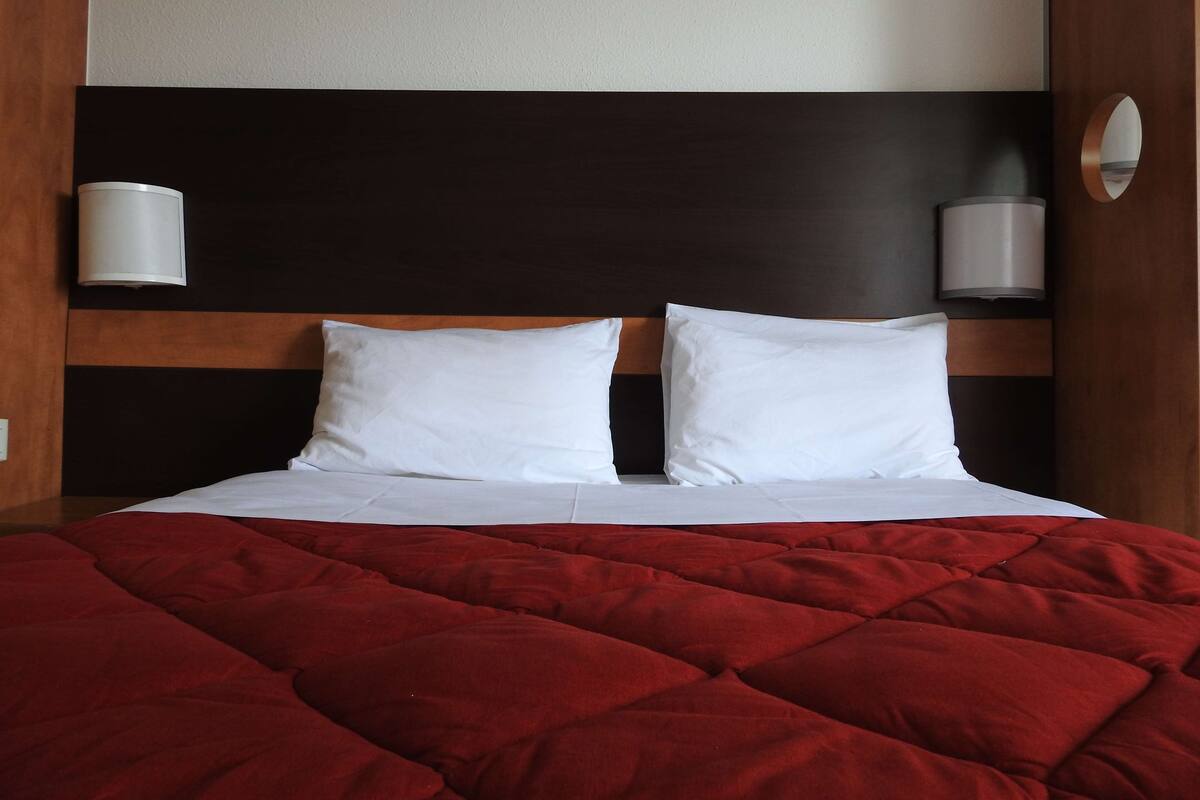 Habitaci ón de hotel con una cama, dos almohadas blancas, una colcha acolchada roja y dos lámparas de pared.