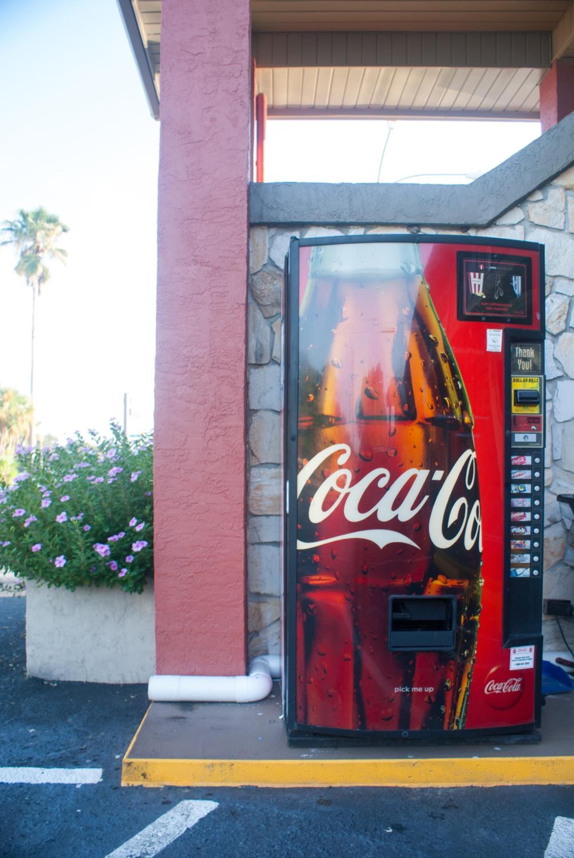 A Coca-Cola vending machine with a red and black design, located next to a building with a stone facade and a red pillar.
