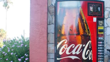 A Coca-Cola vending machine with a red and black design, located next to a building with a stone facade and a red pillar.