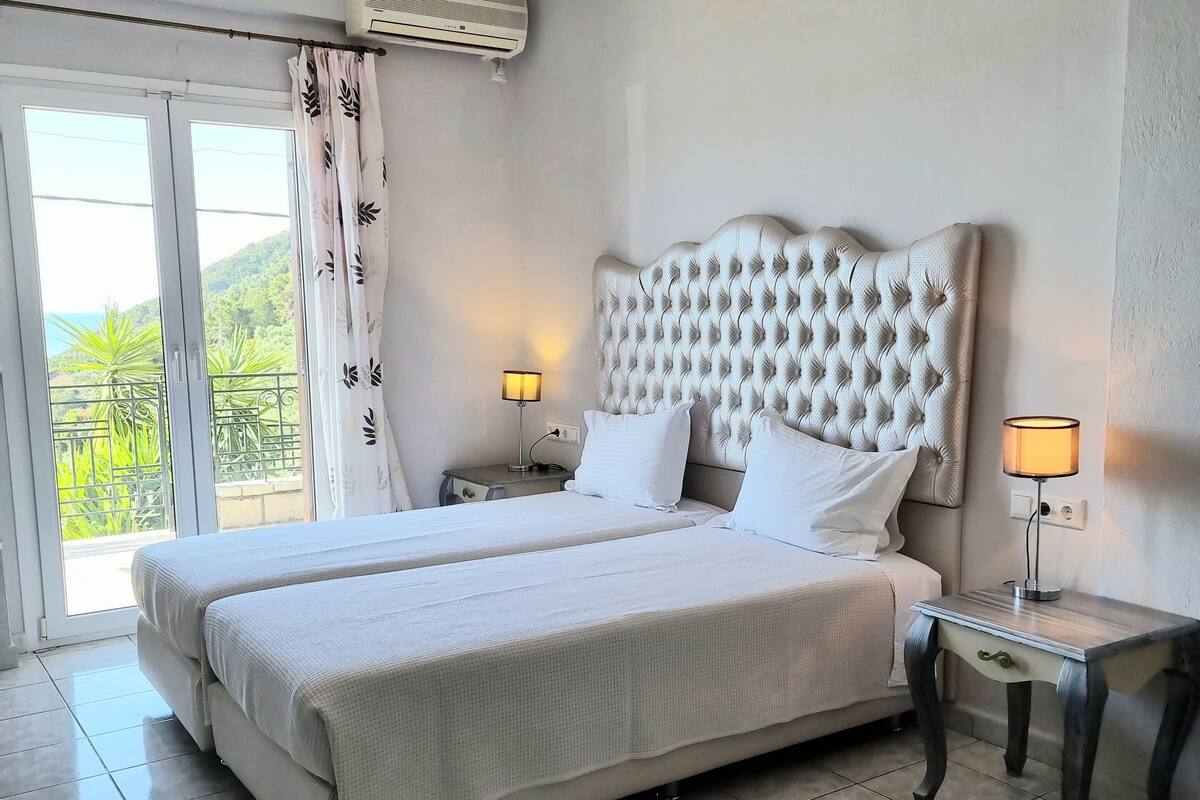 A hotel room with a large bed, two bedside tables, a ceiling light, an air conditioning unit, and a view of greenery through the window.