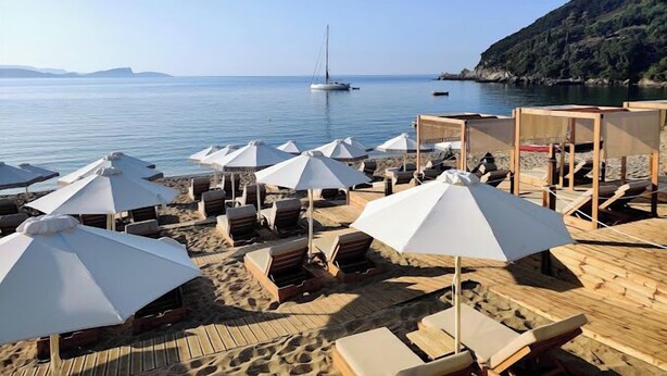 A beach with wooden deck chairs and white umbrellas, a sailboat in the water, and a rocky hillside in the background. - Enjoy Lichnos Bay Village, Camping, Hotel & Apartments (Parga)
