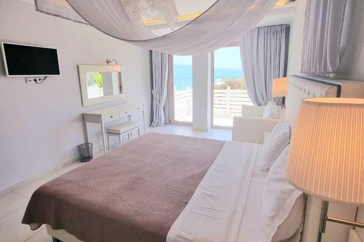 A bedroom with a large bed, a bedside table, a TV, a mirror, and a view of the sea.