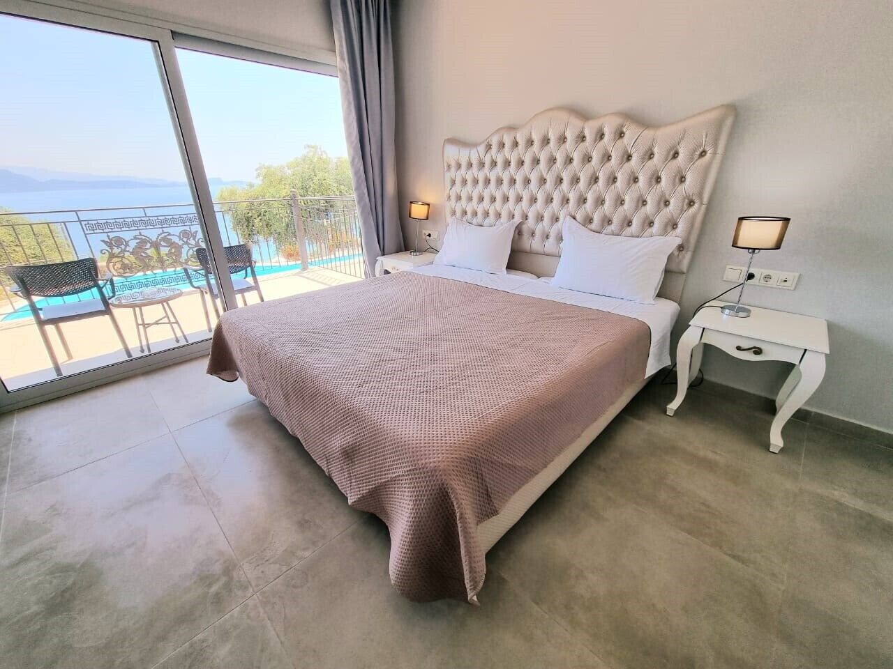 Une chambre dotée d’un grand lit, d’une table de chevet et offrant une vue sur un balcon aménagé avec du mobilier d’extérieur.