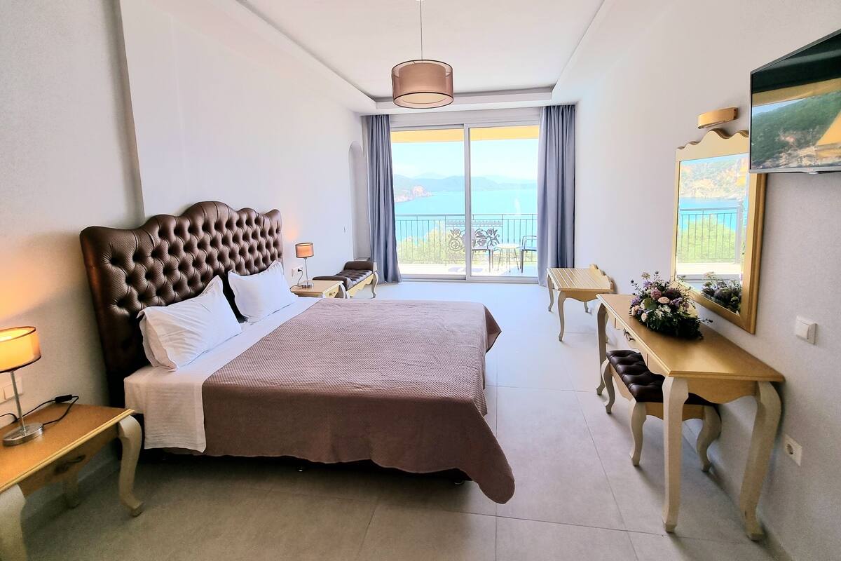 A hotel room with a large bed, a desk, a chair, a TV, and a balcony with a view.