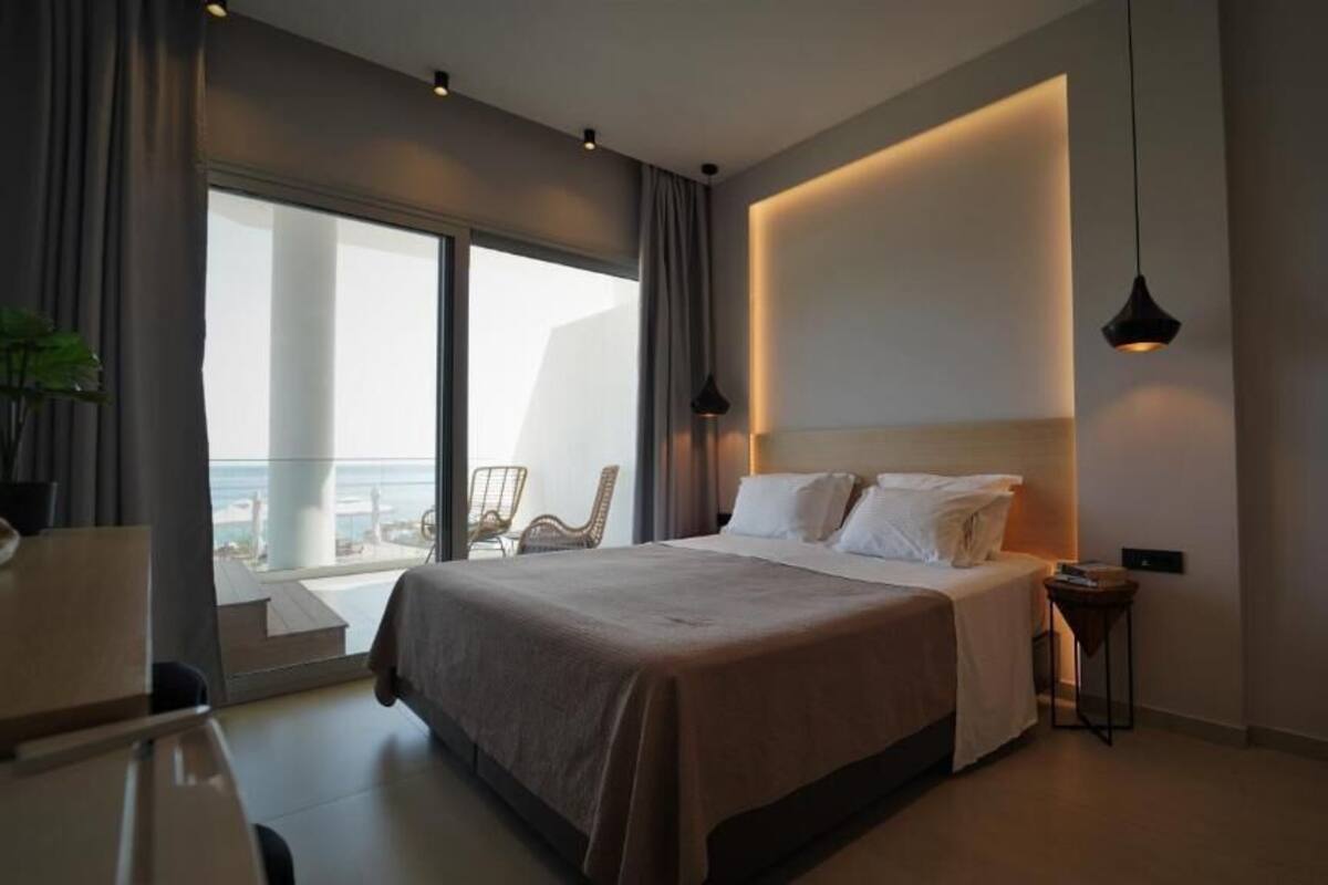 A modern hotel room with a large bed, a bedside table, a chair, and a view of the ocean through large windows.