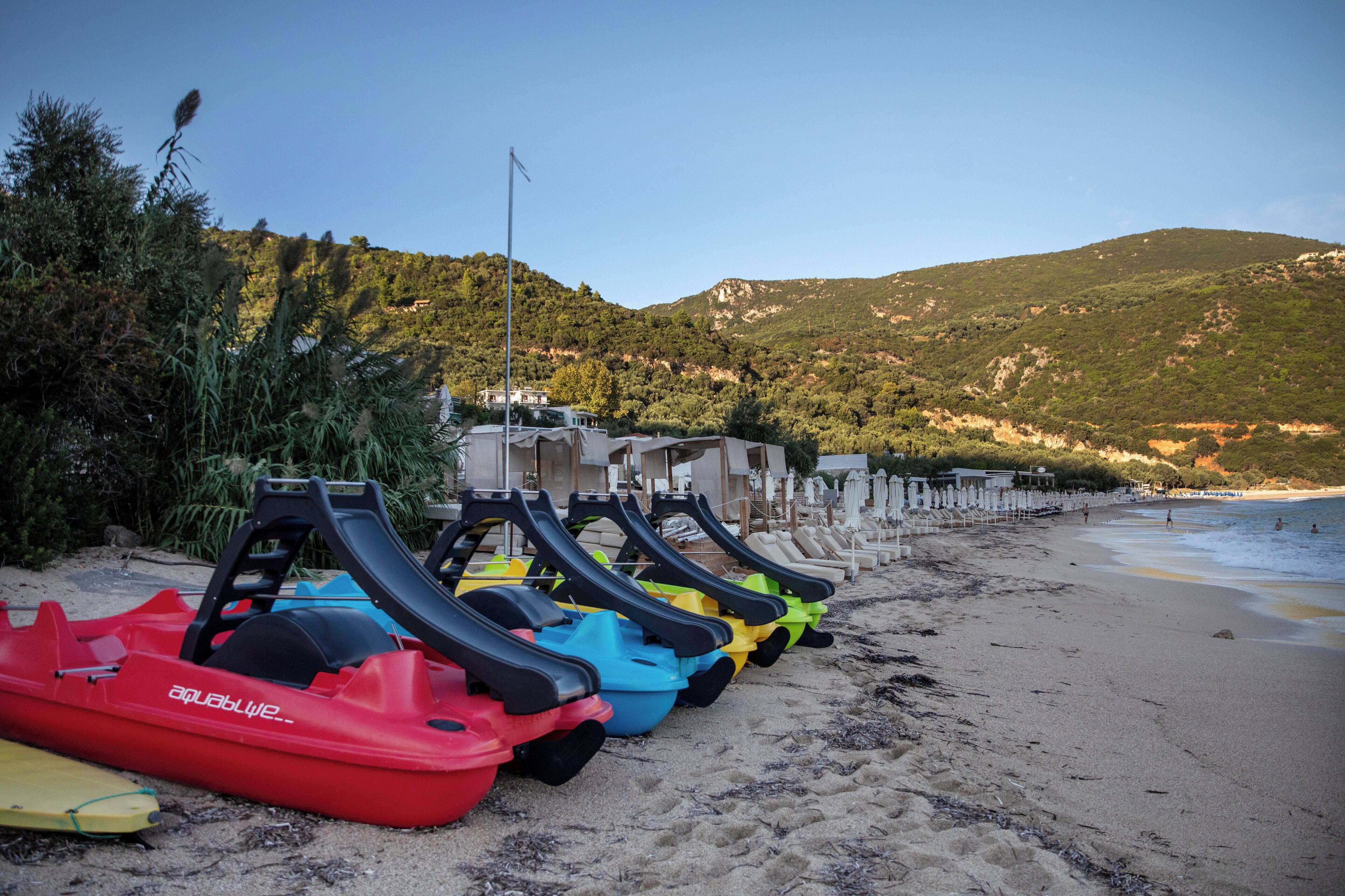 Une rangée de bateaux de différentes couleurs sur une plage de sable, avec en arrière-plan des collines et un ciel dégagé.