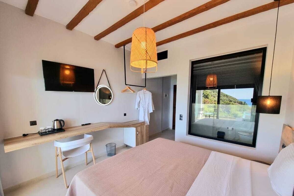 A modern bedroom with a large bed, a wooden desk, a hanging light fixture, and a view of the outdoors through a large window.