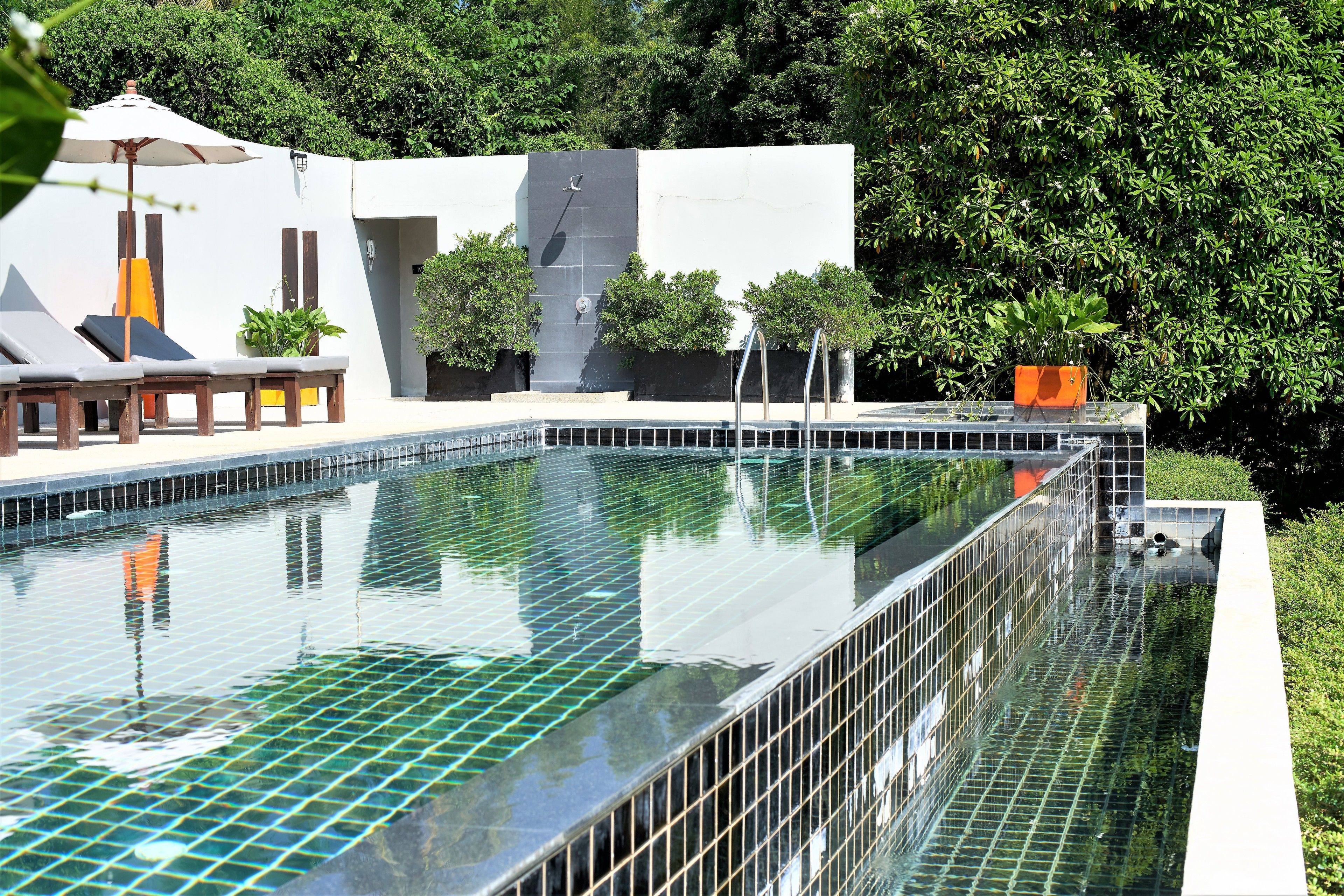 A rectangular infinity pool with a tiled edge, surrounded by a white building with a modern design, including a covered seating area with lounge chairs and a potted plant.