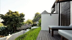 Una terraza de madera con un banco, rodeada de abundante vegetación y un muro blanco.