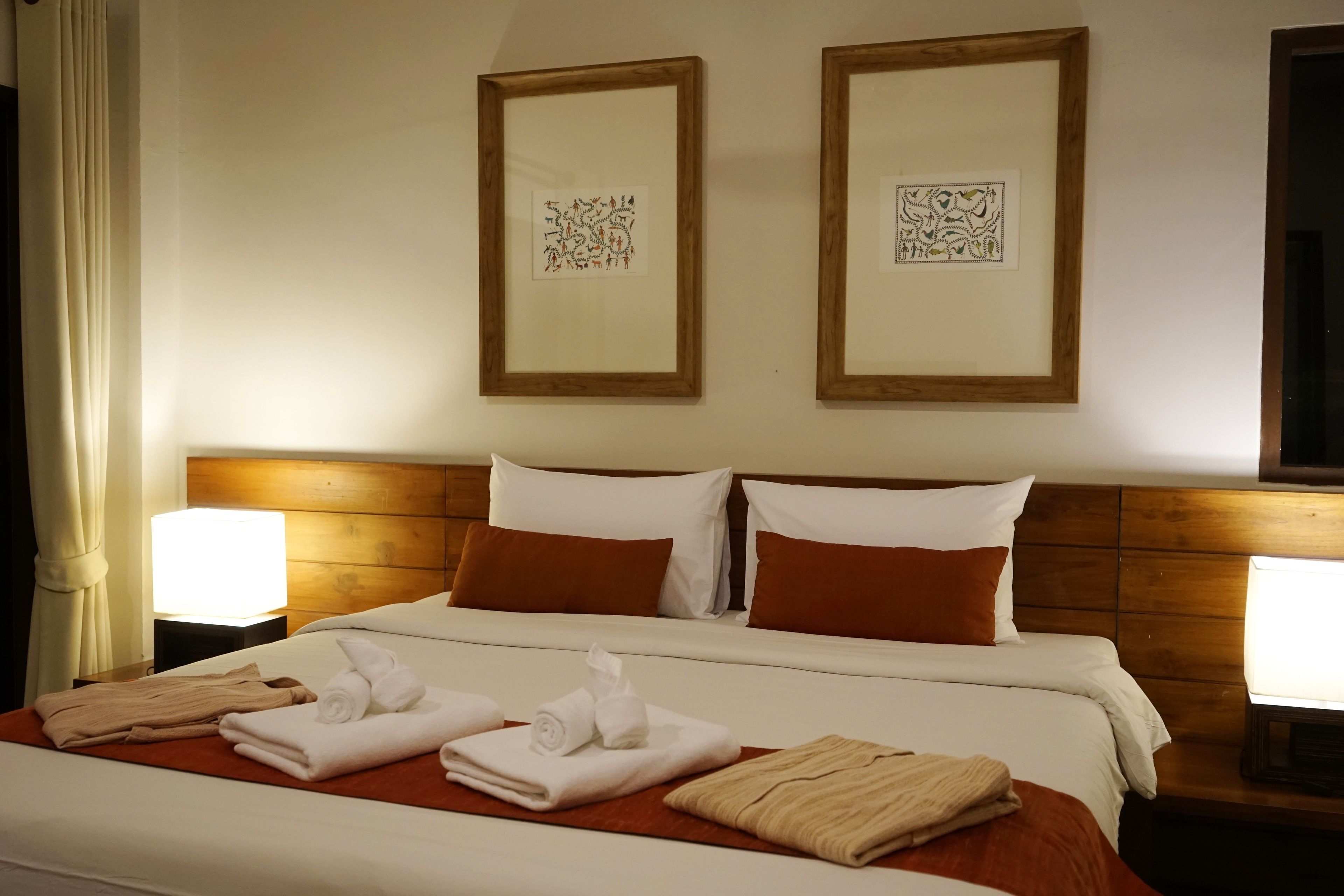 A hotel room with a neatly made bed, two framed artworks on the wall, and a nightstand with a lamp.