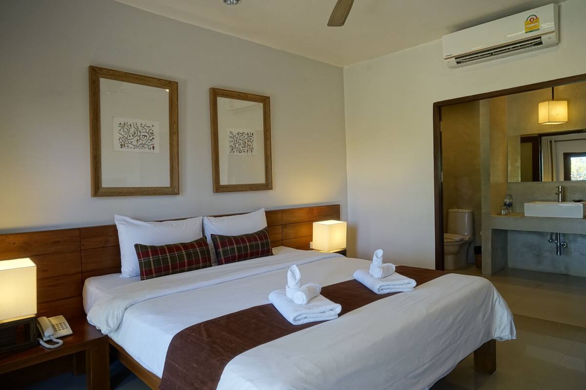 Quarto de hotel com duas camas, cabeceira de madeira, telefone ao lado da cama, ar condicionado fixado na parede e banheiro com pia e espelho.
