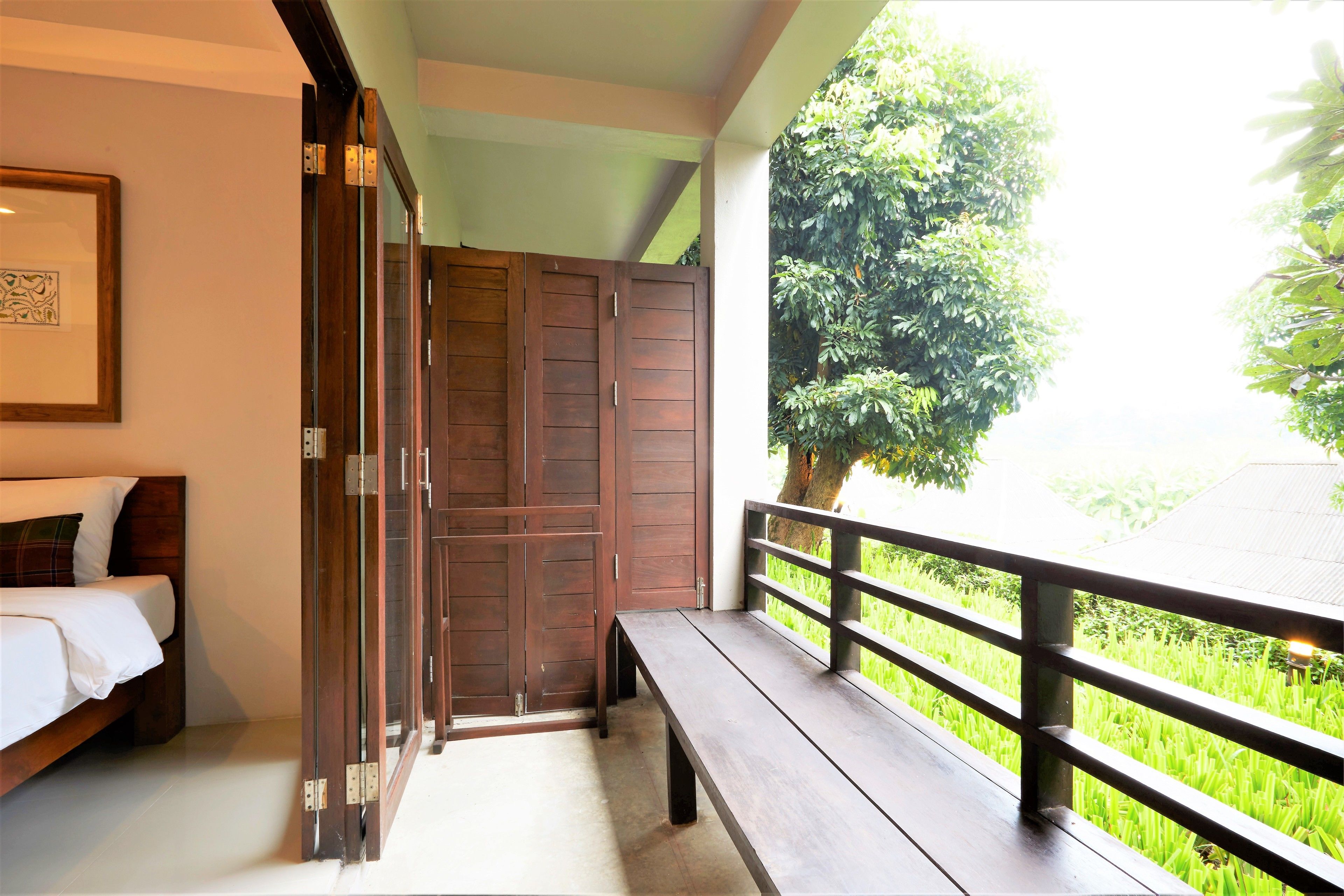 A room with a bed, a bench, and a sliding door leading to a balcony with a view of greenery.
