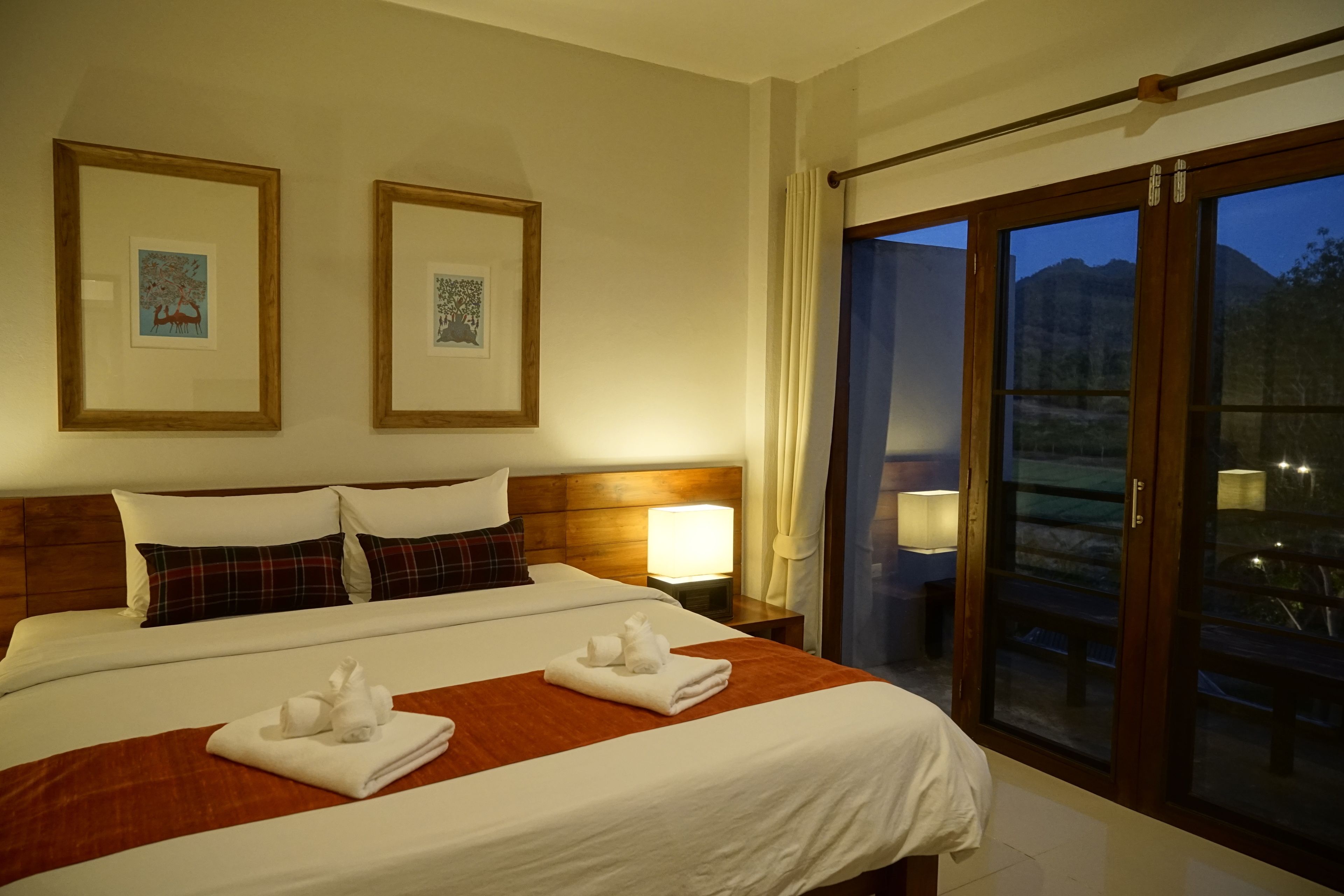 A hotel room with a large bed, two framed pictures on the wall, a nightstand with a lamp, and a sliding glass door leading to an outdoor area.