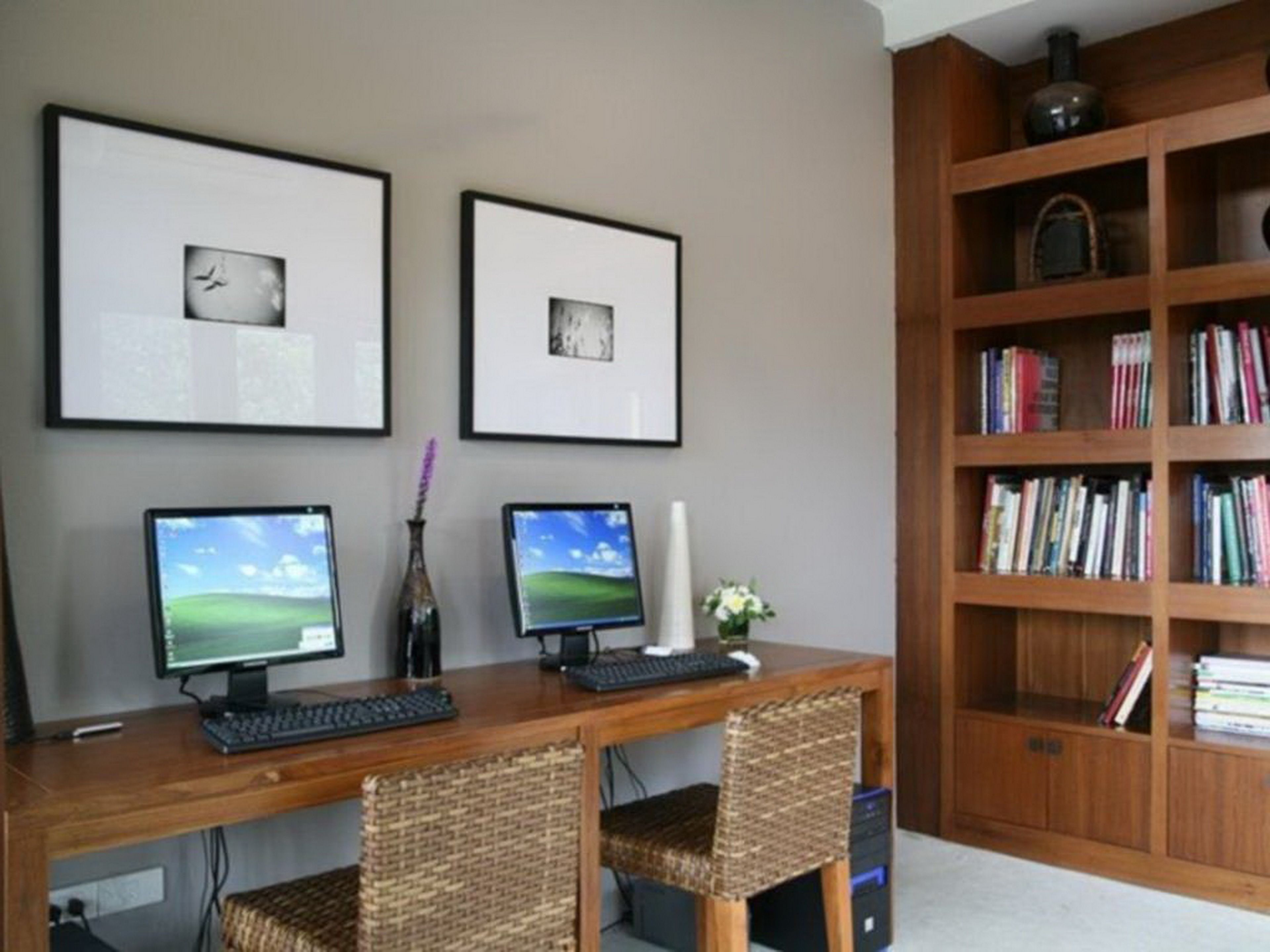 Una oficina en casa con dos computadoras de escritorio, una estantería y fotografías enmarcadas en la pared.