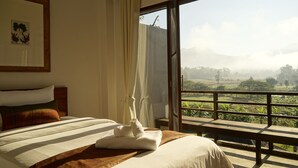 Habitación de hotel con cama, vista a un paisaje verde a través de un balcón y un cuadro en la pared.