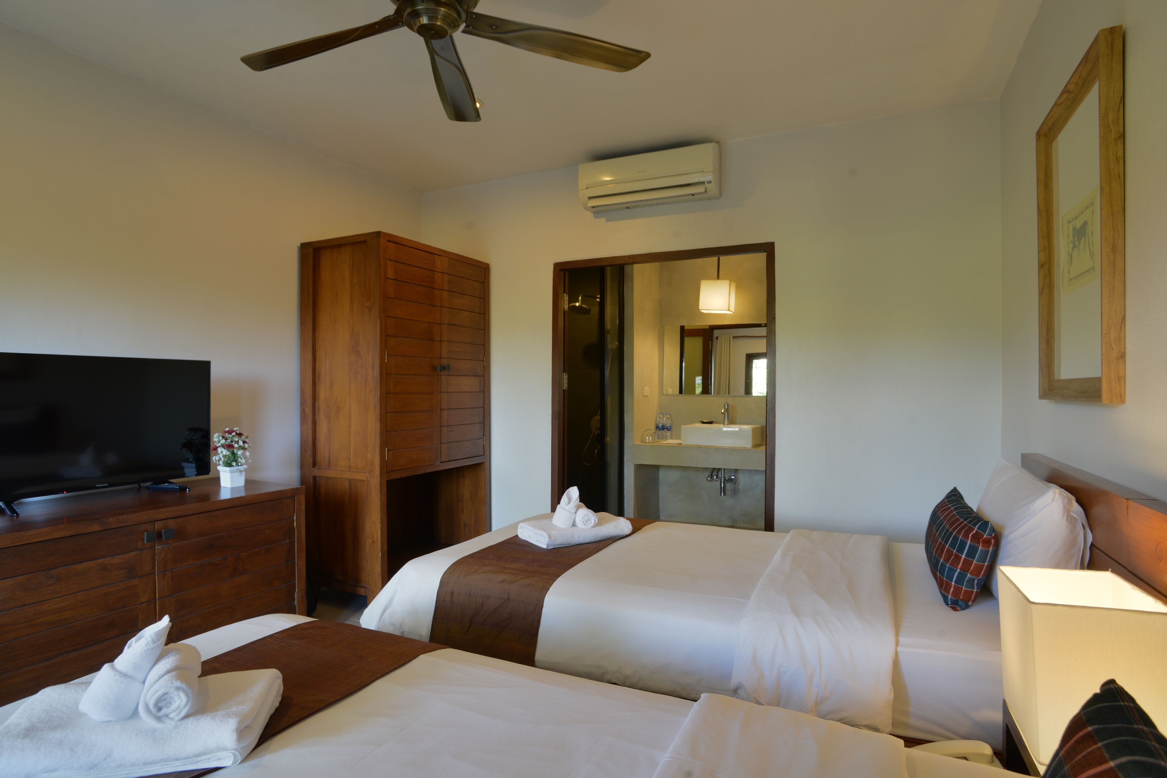 A hotel room with two beds, a TV, a wooden wardrobe, a ceiling fan, and a bathroom visible through an open door.