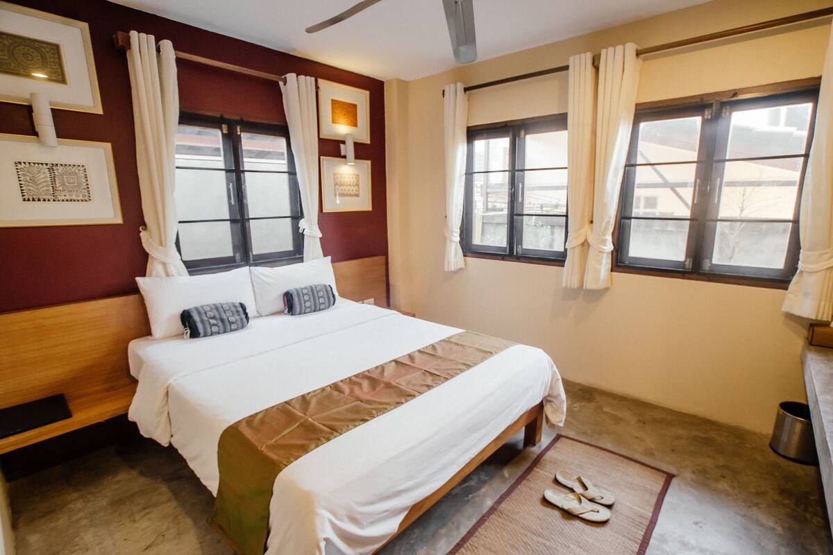 Habitación de hotel con cama, mesita de noche, ventana con cortinas y un cuadro en la pared.