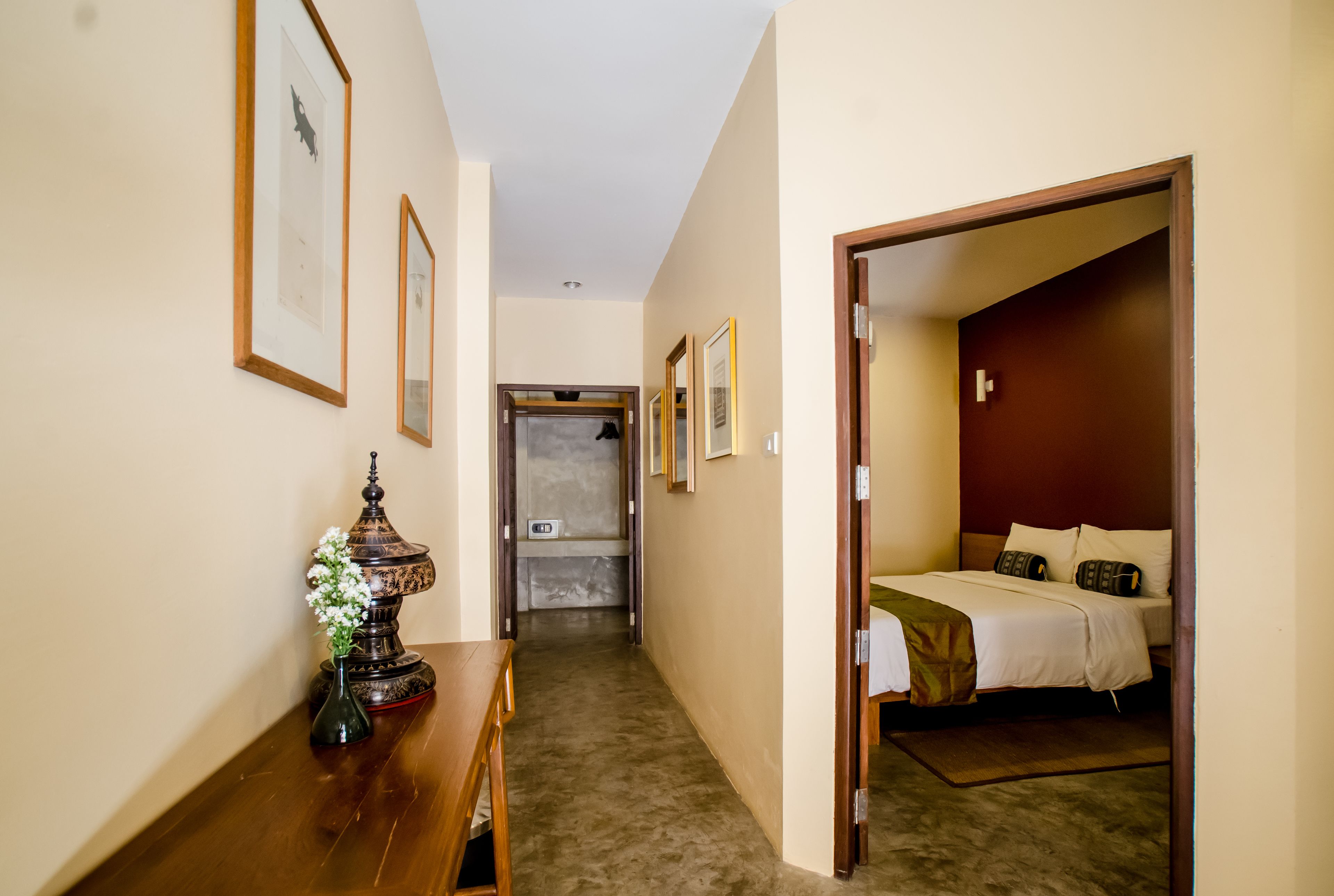 A hotel hallway with framed artwork on the walls, a wooden bench, and a bed in the adjacent room.
