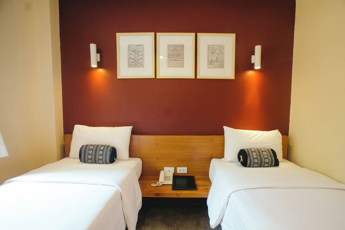 Habitación de hotel con dos camas, cabecera de madera, mesita de noche con un teléfono y una tableta, y tres cuadros en la pared.