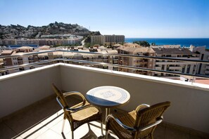 A balcony with wicker chairs and a small table, overlooking a coastal town and the sea.