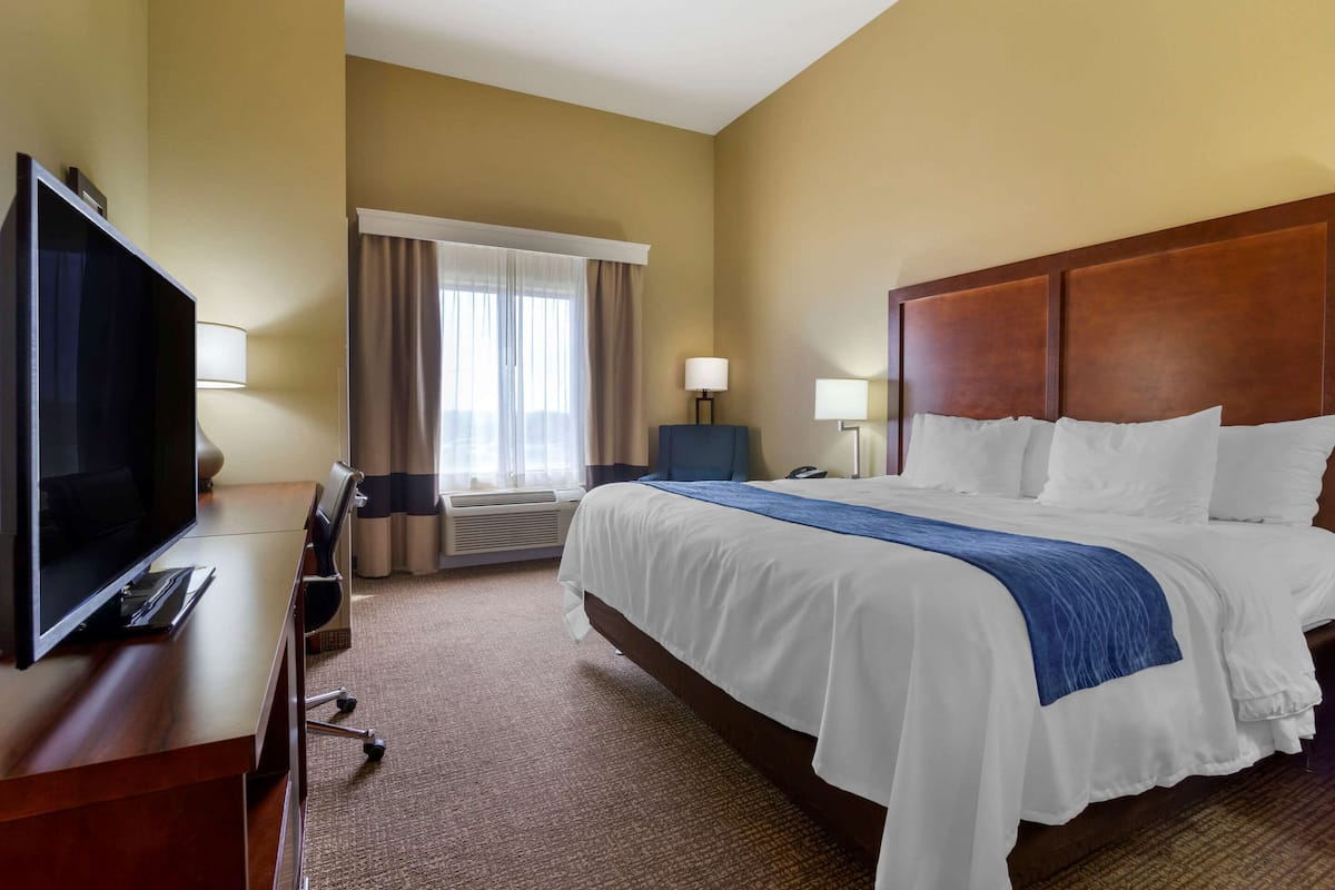 A hotel room with a large bed, a desk with a TV, a lamp, and a window with curtains.
