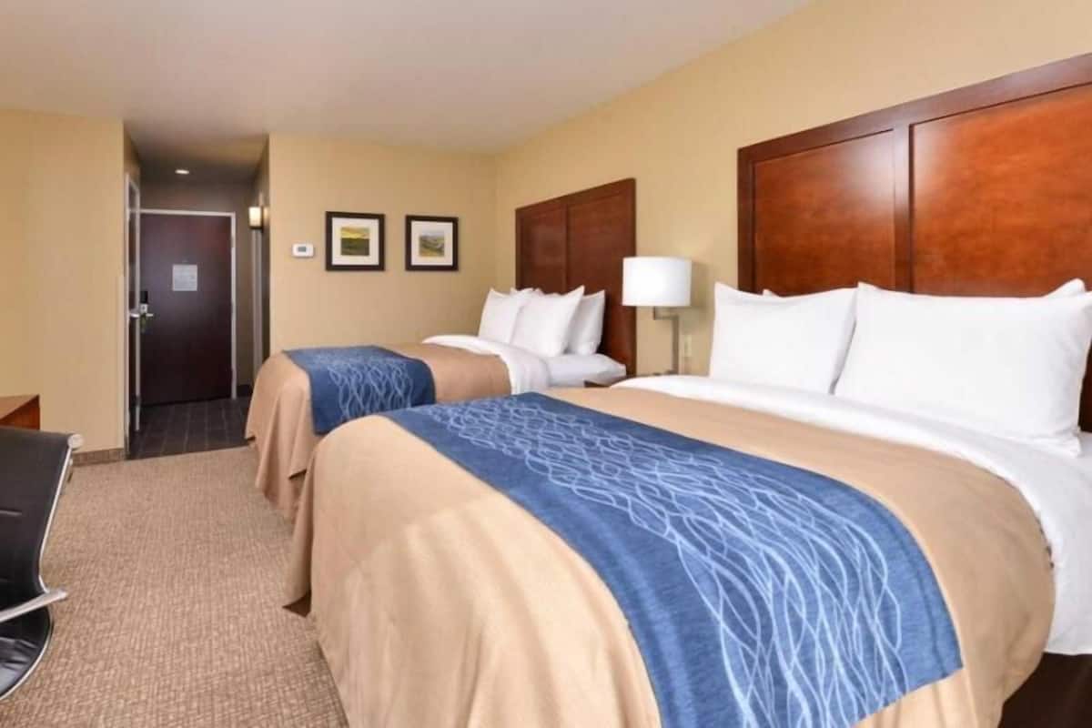 A hotel room with two beds, a desk, a chair, and a door leading to a hallway.