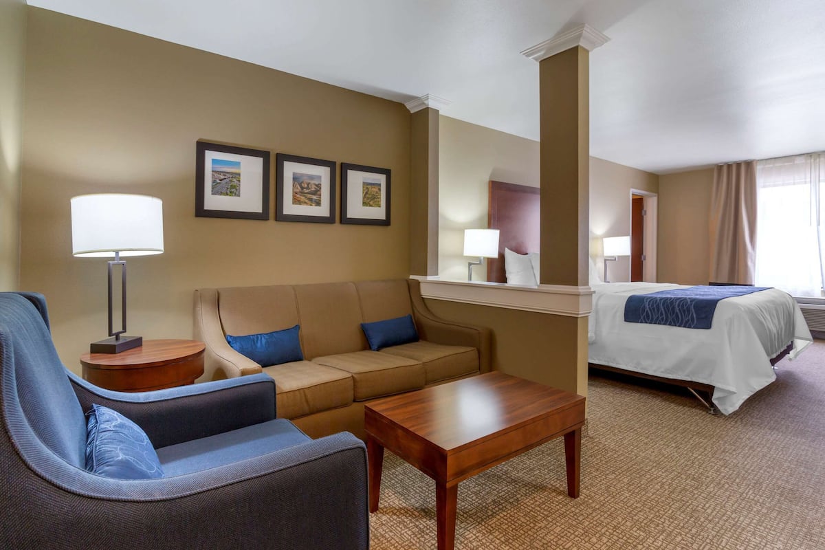 A hotel room with a sofa, armchair, bed, and a small table. There are framed pictures on the wall and a lamp on the side table.