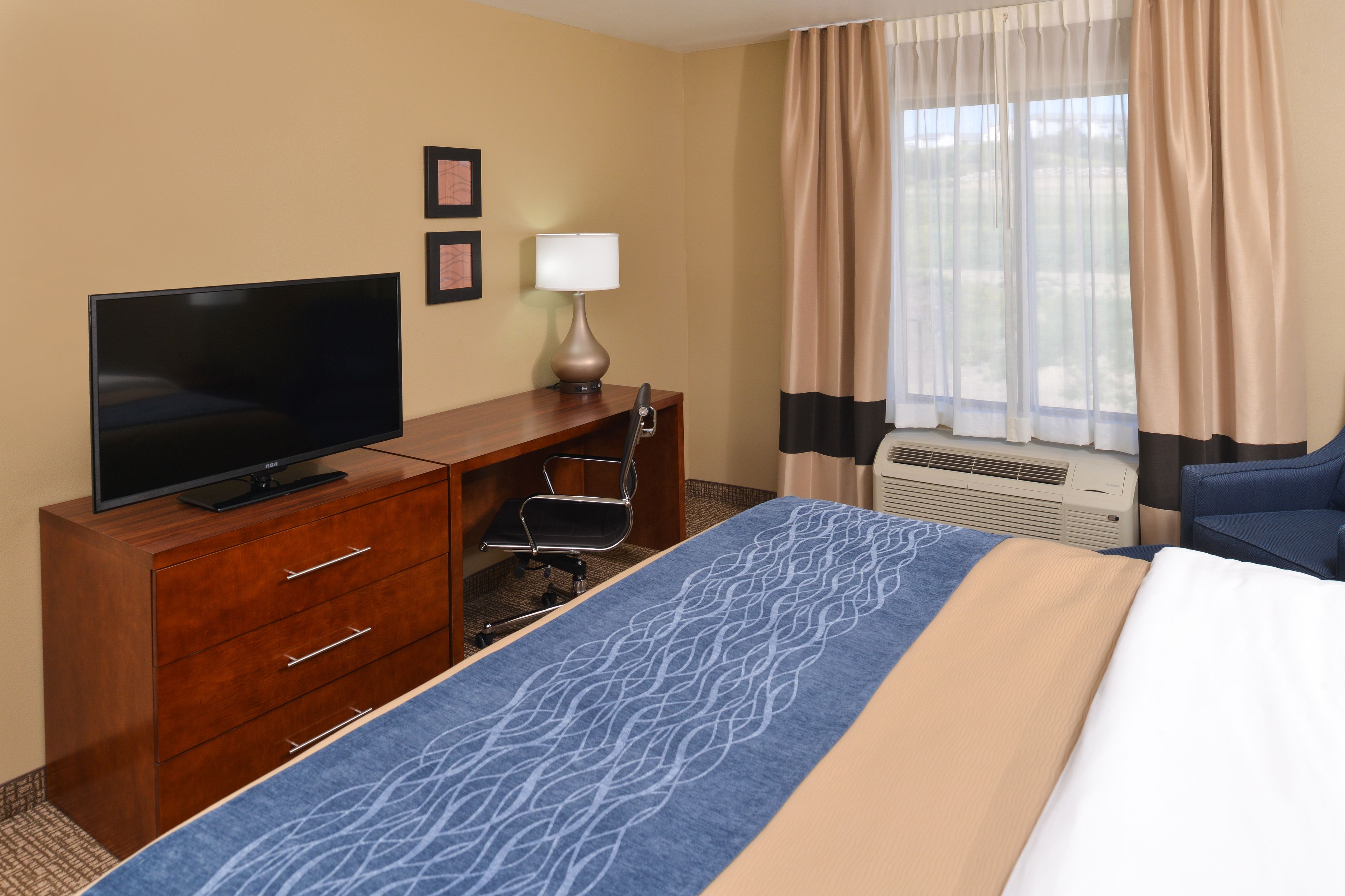 A hotel room with a bed, a TV on a wooden dresser, a desk with a chair, a lamp, and a window with curtains.
