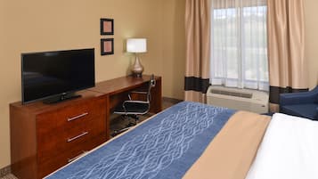 A hotel room with a bed, a TV on a wooden dresser, a desk with a chair, a lamp, and a window with curtains.