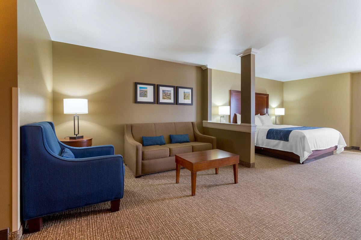 A hotel room with a blue armchair, a sofa, a wooden coffee table, a bed with a white comforter, and three framed pictures on the wall.