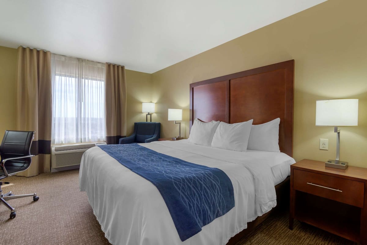A hotel room with a large bed, two bedside lamps, a desk with a lamp, a chair, and a window with curtains.