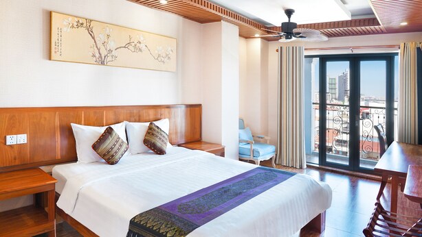 Ein Hotelzimmer mit einem großen Bett, einem Holz-Kopfteil, einem Deckenventilator, einem Balkon mit Blick auf die Stadt und einem traditionellen Kunstwerk an der Wand.