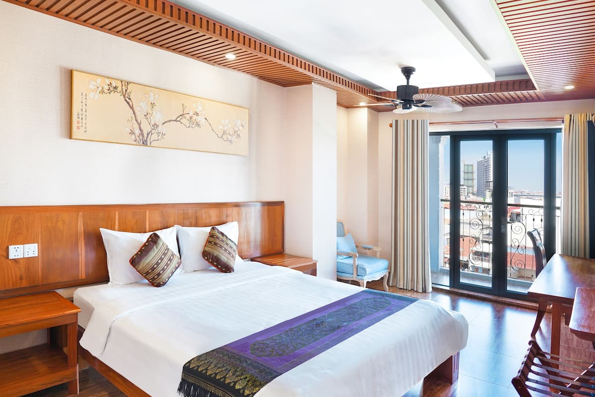 Habitación de hotel con una cama grande, cabecera de madera, ventilador de techo, balcón con vistas a la ciudad y una obra de arte tradicional en la pared.