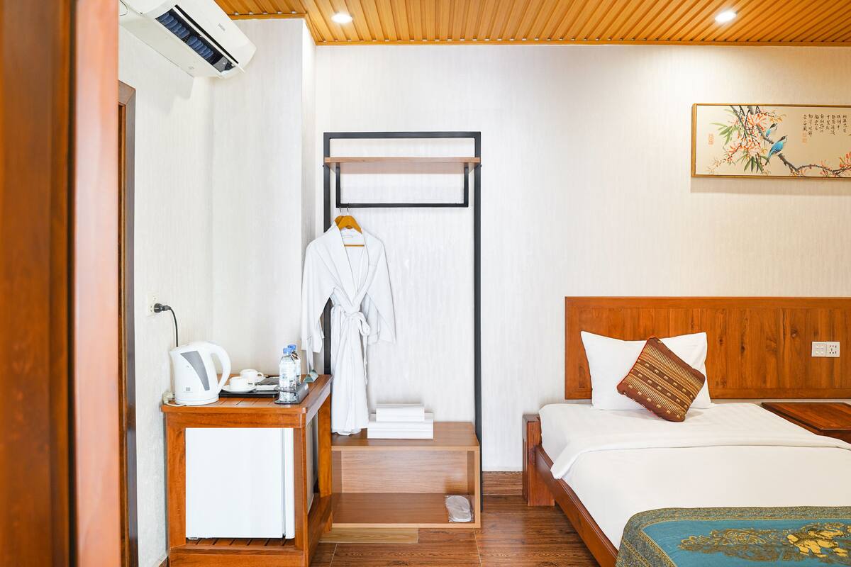 Una habitación de hotel con una cama de madera, una mesita de noche con una tetera, una bata colgada en un perchero y un estante en la pared.