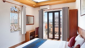 Ein Hotelzimmer mit Bett, Schreibtisch, Stuhl, einem Fenster mit Vorhängen und Blick auf die Stadt durch eine Glastür.