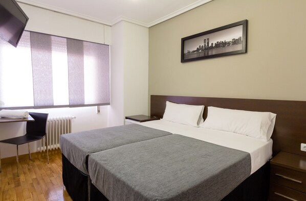 A hotel room with two beds, a desk, a chair, a radiator, and a cityscape picture on the wall.