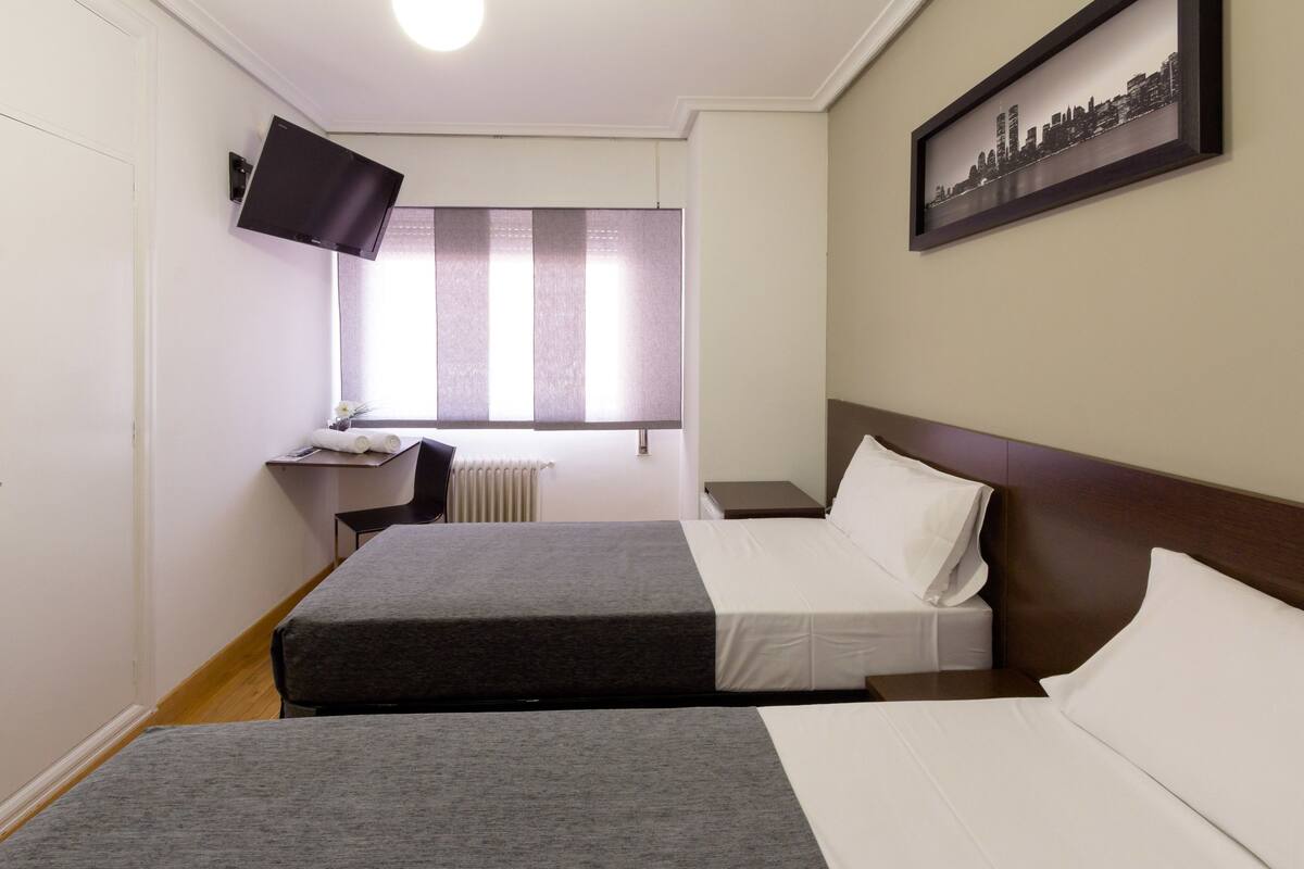 A hotel room with two beds, a desk, a chair, a window with blinds, a flat-screen TV mounted on the wall, and a framed cityscape picture.