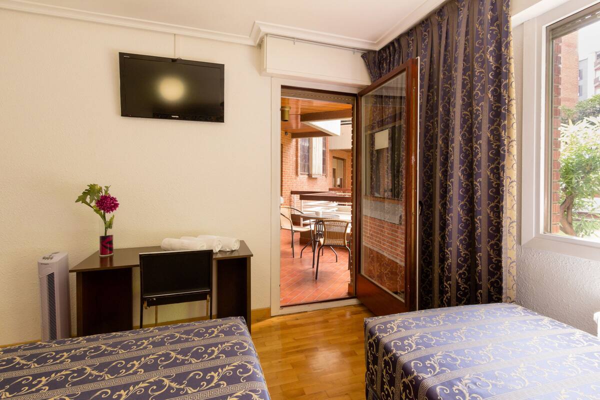 A hotel room with two beds, a desk, a TV, and a sliding glass door leading to a dining area.