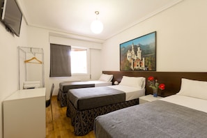A hotel room with two beds, a TV, a window with curtains, a painting on the wall, and a small table with a vase of flowers.