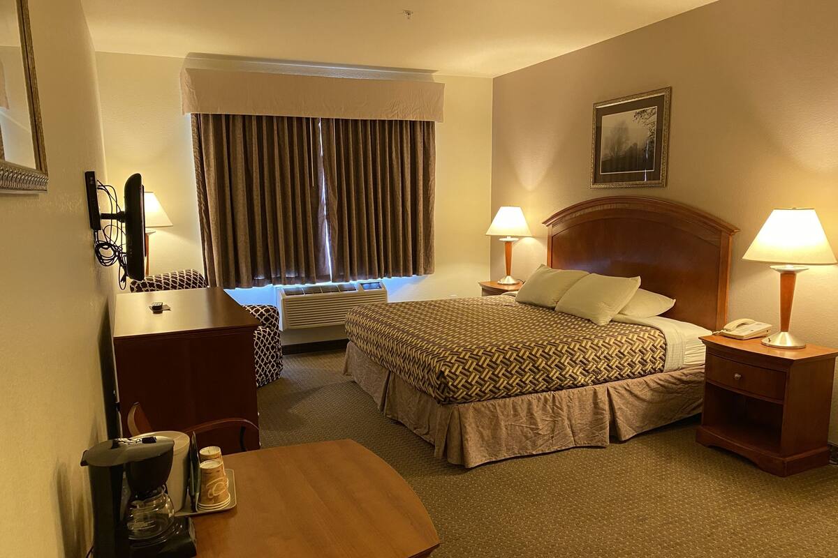 A hotel room with a bed, bedside lamps, a desk with a coffee maker, a chair, a dresser, and a window with curtains.