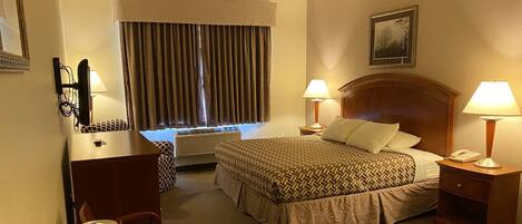 A hotel room with a bed, bedside lamps, a desk with a coffee maker, a chair, a dresser, and a window with curtains.