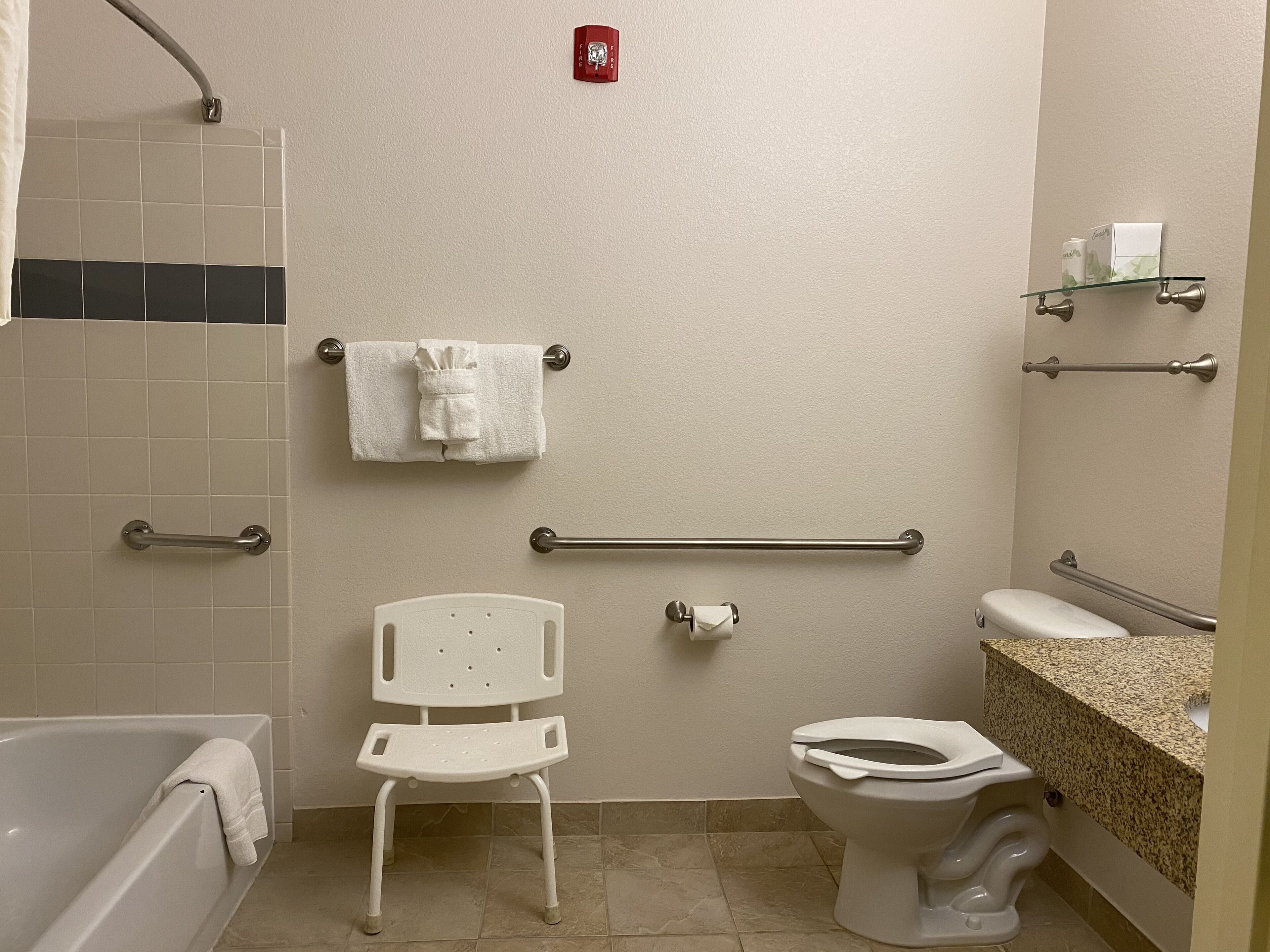 A bathroom with a bathtub, toilet, and a shower area. There are grab bars installed for safety, a towel rack, and a glass shelf with toiletries.