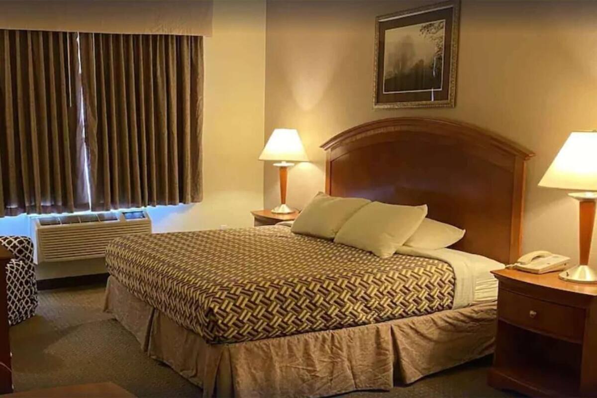 A hotel room with a large bed, two bedside lamps, a nightstand with a phone, a window with curtains, and a framed picture on the wall.
