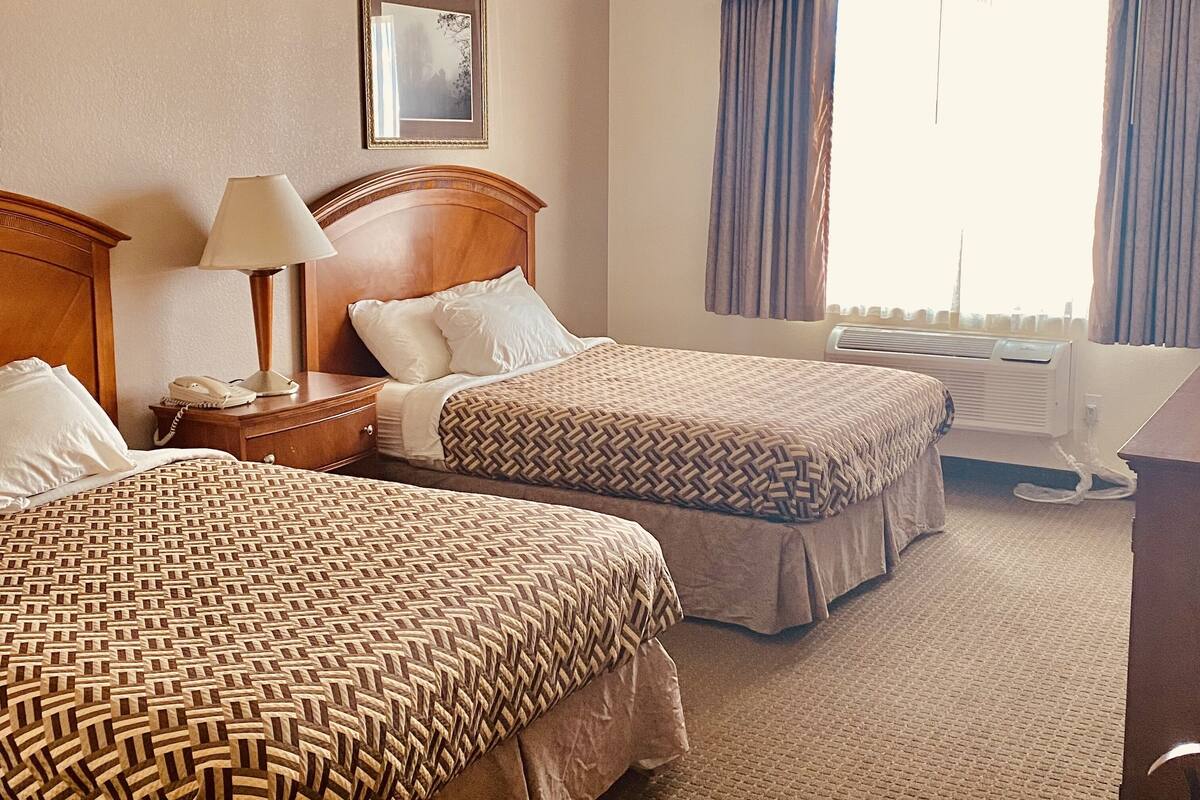 A hotel room with two beds, a bedside table with a lamp, a phone, and a framed picture on the wall.