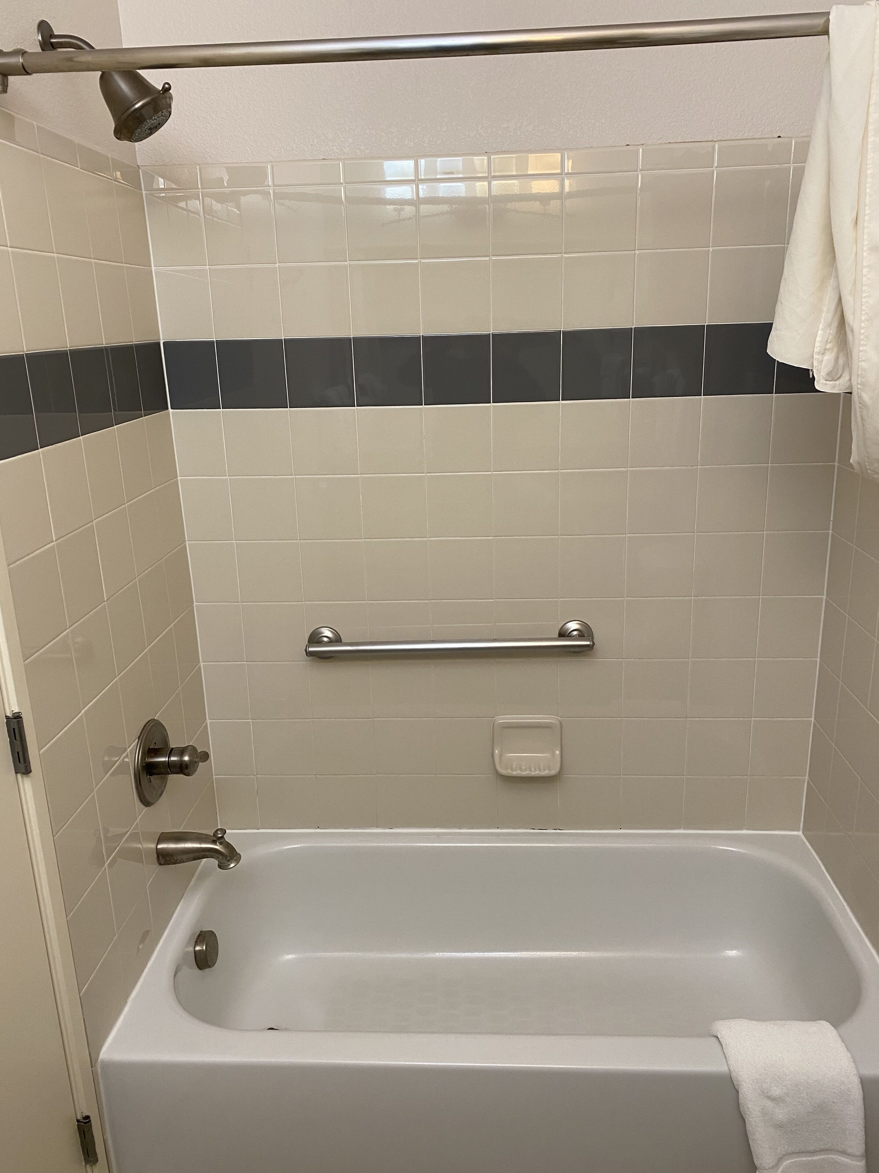 A standard hotel bathroom with a bathtub, a showerhead, a handrail, and a towel hanging on the wall.