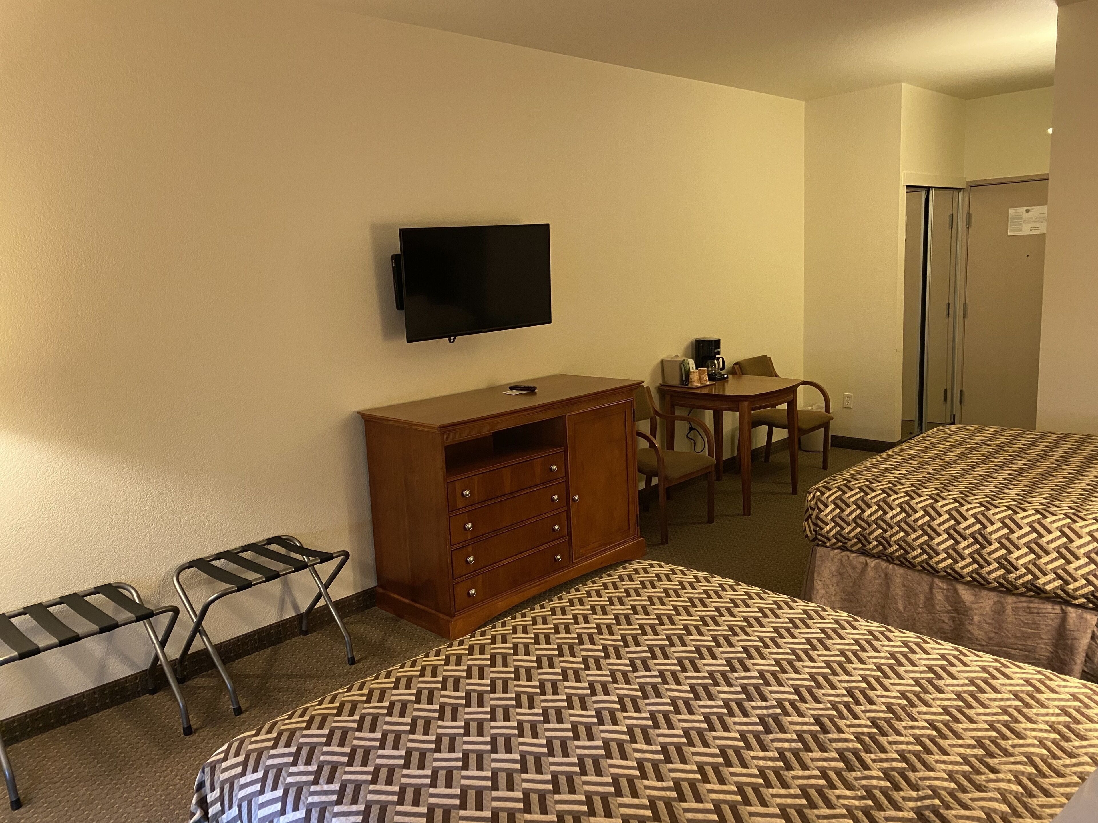 A hotel room with two beds, a TV, a wooden dresser, a desk with a chair, and a small table with a coffee maker.