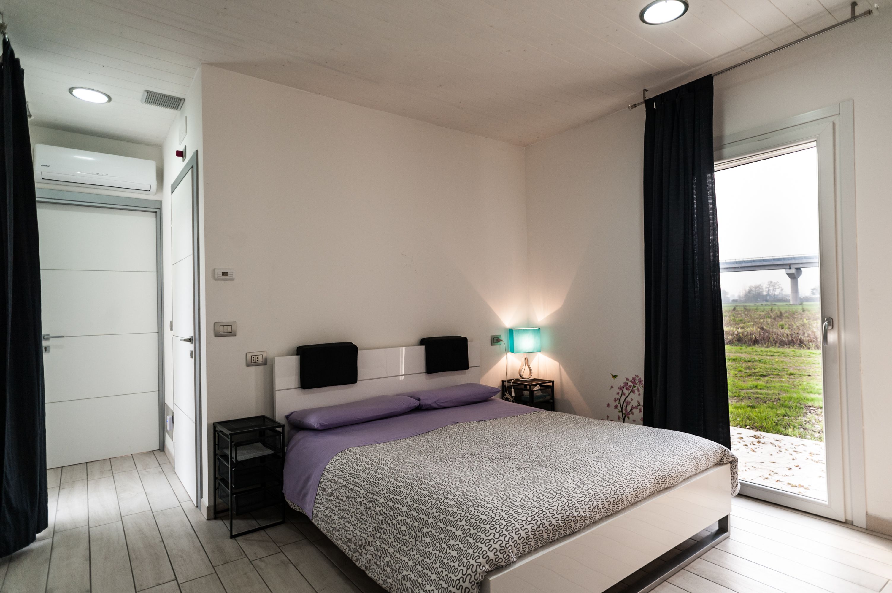 A bedroom with a bed, a bedside table, a lamp, and a view of the outdoors.