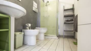 A modern bathroom with a large white sink, two toilets, a glass shower enclosure, and a green laundry basket.