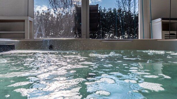 Una piscina con agua cristalina, vista a árboles y montañas a través de un ventanal, y una escultura de metal en la pared.